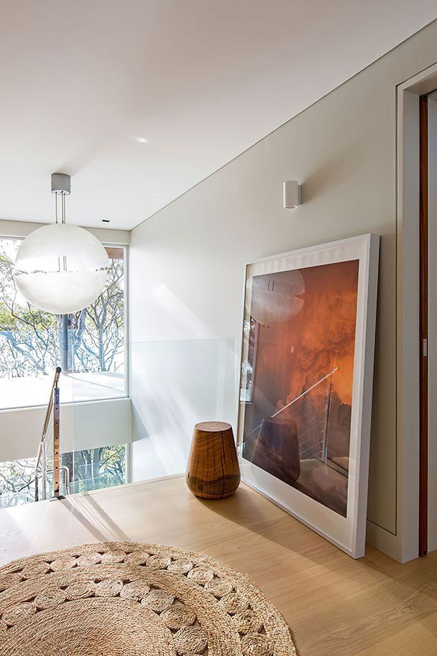 Bright contemporary upper-level landing or hallway with pale timber flooring, white walls and ceiling, a glass balustrade overlooking a stairwell, and large windows. A sculptural spherical pendant light, framed artwork, wooden vessel, and round woven rug add warm natural accents.