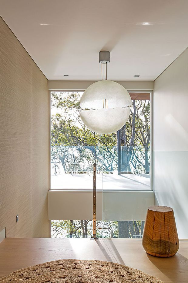 Bright contemporary interior viewed across a timber dining table toward a full-height glazed opening and stair or landing area. A large spherical pendant light hangs centrally, with a warm textured wall finish on the left, pale walls, glass balustrading, and a sculptural timber table lamp. Trees are visible outside through the glazing, and a woven rug occupies the foreground.