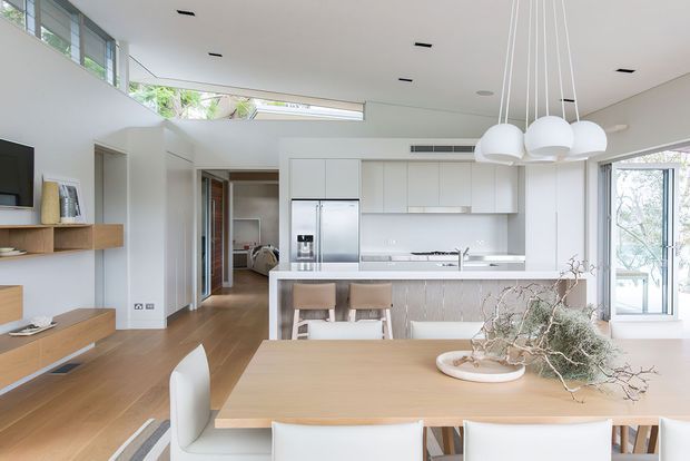 Bright open-plan contemporary kitchen and dining area with white built-in cabinetry, stainless-steel refrigerator, pale timber island and dining table, integrated living-room joinery, expansive glazing, and sculptural white pendant lights. The space has a clean, airy, minimalist appearance with warm timber flooring and furnishings.