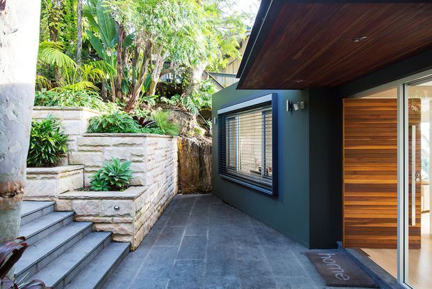 Modern residential entry courtyard with dark charcoal exterior walls, a timber-lined soffit, and a timber front door with horizontal glazing. The entry is approached by broad grey tiled steps and paving, alongside layered sandstone garden retaining walls and lush tropical planting.