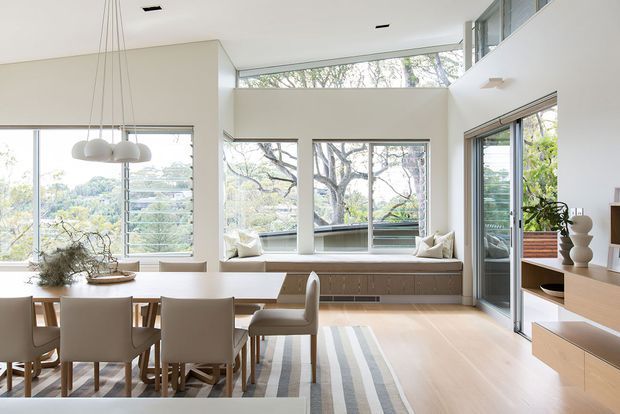 Bright, airy open-plan dining and living space with a large dining table, upholstered chairs, built-in window bench seating, pale timber flooring, extensive glazing, and a double-height ceiling. Minimal built-in cabinetry and a sculptural pendant light create a refined contemporary interior with views to leafy surroundings.