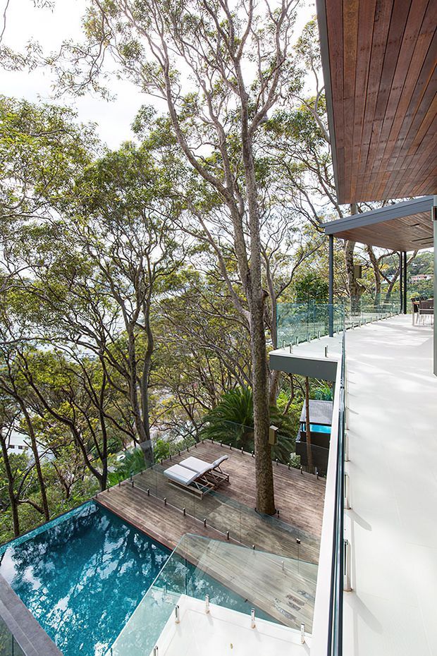 Elevated view of a modern multi-level home integrated into a heavily wooded sloping site, with a turquoise swimming pool, timber pool deck, glass balustrades, and expansive white outdoor terraces. Timber soffits and clean-lined construction frame the landscaped outdoor living areas.