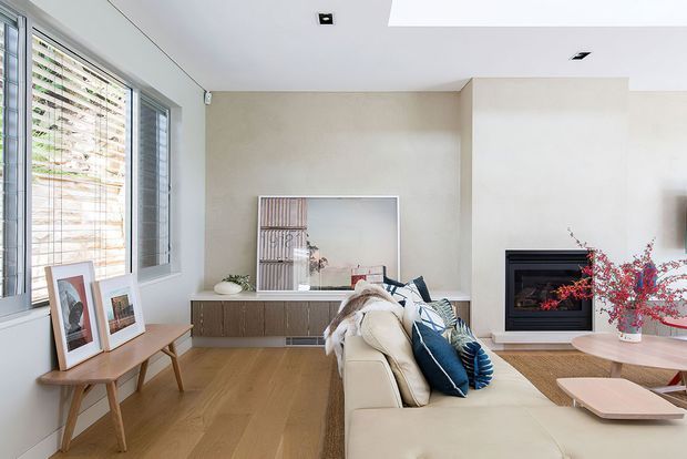 Bright contemporary living room with a cream sectional sofa, timber flooring, a built-in fireplace, low timber media cabinetry, large windows with blinds, and layered coffee tables. Neutral finishes are accented by navy cushions and red foliage.