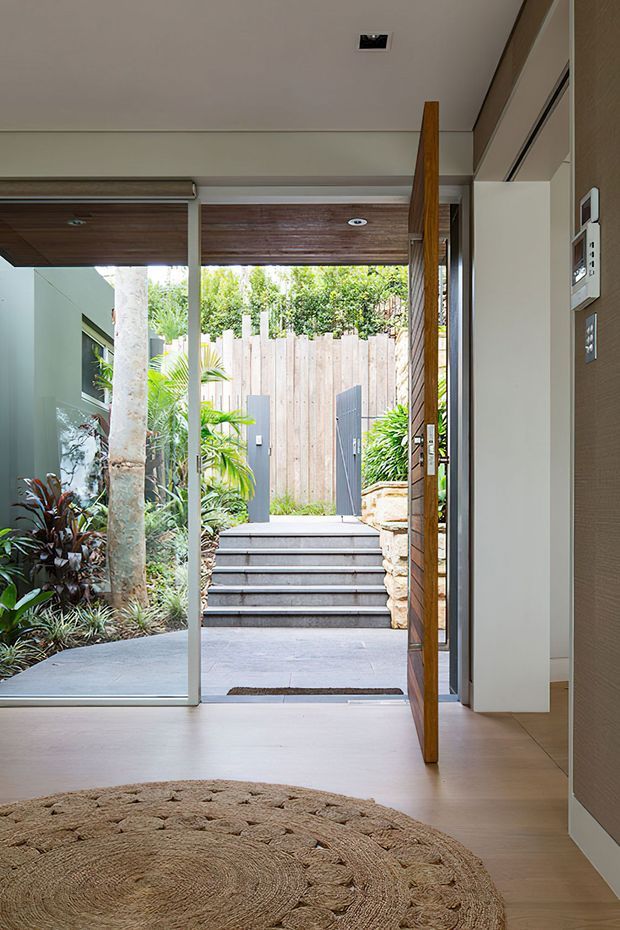 A bright, modern entryway viewed from inside toward a landscaped courtyard. The space features pale timber flooring, a large frameless glass opening, a tall timber pivot door, white walls, recessed ceiling lights, and a textured round natural-fibre rug. Outside, broad steps lead to a timber privacy fence and planted garden, with exposed stone retaining elements at the right.