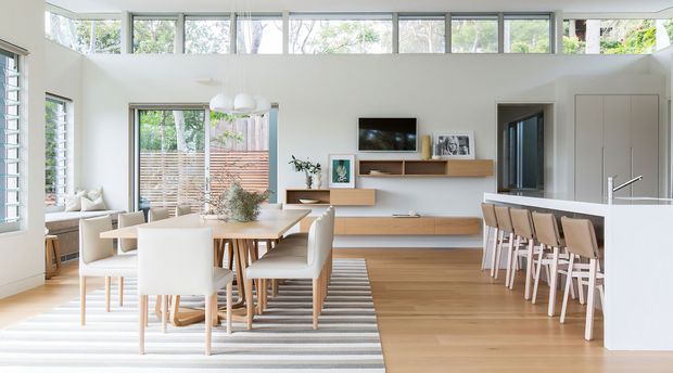Bright open-plan contemporary living, dining and kitchen area with extensive clerestory glazing, white walls, pale timber flooring, a timber dining table, built-in timber media shelving, and a white kitchen island with integrated seating. The space has abundant natural light and a clean, airy feel.