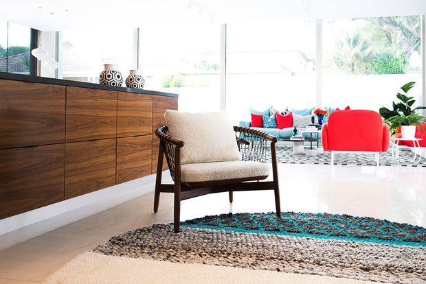 Bright, airy mid-century-inspired living room with a walnut-toned built-in sideboard, woven lounge chair, vivid red and turquoise seating, patterned rug, indoor plants, and expansive floor-to-ceiling glazing.