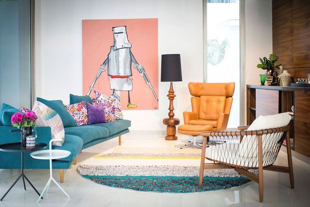 A bright, eclectic living room with a teal sofa layered with colourful patterned cushions, an orange leather armchair, a wire-frame lounge chair, a sculptural floor lamp, and overlapping patterned rugs. The room features large glazing, white walls and polished pale flooring, with dark timber built-in cabinetry along one side and colourful artwork as a focal point.