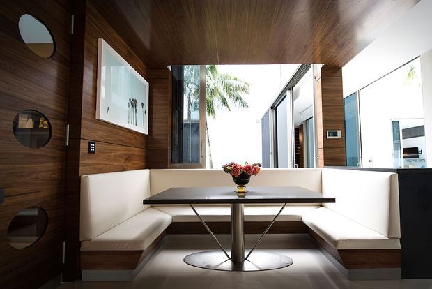A compact, refined dining nook with built-in white upholstered banquette seating arranged around a dark rectangular table with a central pedestal base. Rich timber wall and ceiling panelling create a warm architectural enclosure, while a large glazed opening brings in daylight and views of palm trees. Circular wall niches, a framed artwork, recessed lighting, and a visible adjacent courtyard or exterior add visual interest.
