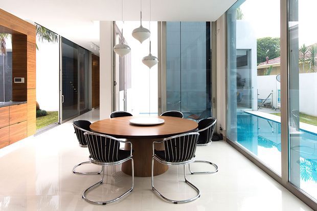 Bright contemporary dining area with a round timber dining table and sculptural black-and-chrome chairs, surrounded by floor-to-ceiling glazing. A swimming pool and landscaped courtyard are visible outside, while timber built-in cabinetry and a wall-mounted TV appear to the left. Multiple pendant lights hang above the table.