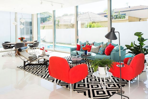 Bright, spacious contemporary living room with floor-to-ceiling glazing overlooking a pool and neighbouring residences. The room features a teal sectional sofa, vivid red lounge chairs and cushions, a black-and-white geometric rug, sculptural pendant lighting, indoor plants, and glossy pale flooring.