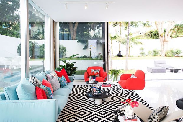 Bright, open-plan living room with floor-to-ceiling glazing overlooking a landscaped courtyard. The room features a pale turquoise sectional sofa, vivid red lounge chairs and cushions, a bold black-and-white geometric rug, a round coffee table, indoor plants, and polished light flooring. White walls and ceiling with discreet track lighting create an airy, contemporary interior.