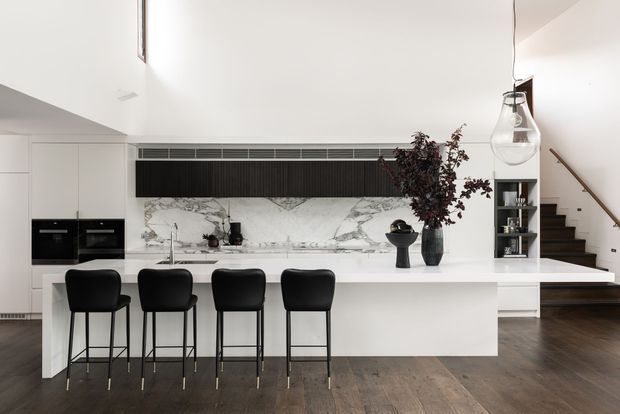 Bright contemporary open-plan kitchen with a large white island and four black bar stools, integrated appliances, a dramatic white-and-grey marble splashback, dark linear feature cabinetry, and dark timber flooring. A sculptural pendant light, open shelving, decorative vessels, and a stairway are visible at the edge of the space.