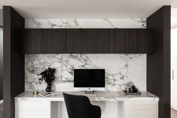 A refined built-in home office workspace with a white marble-look feature wall and desktop, white drawers, dark timber side panels, and a dark fluted overhead cabinet. A computer, black task chair, books, and decorative objects complete the compact workstation.