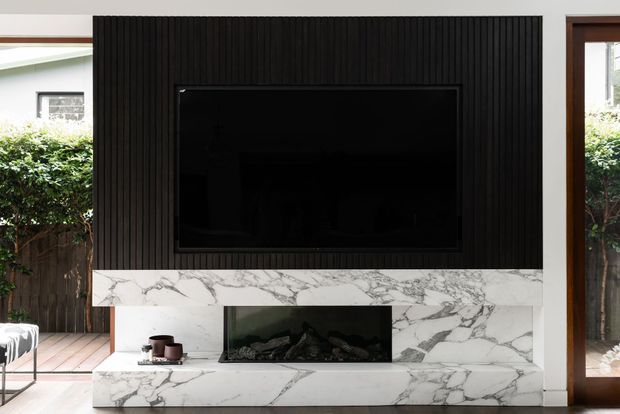 Contemporary living-room feature wall with a large black television set into vertically grooved dark timber panelling. A broad white marble fireplace surround and hearth with prominent grey veining frame a black gas fireplace, flanked by timber-framed glazing and views to greenery outside.