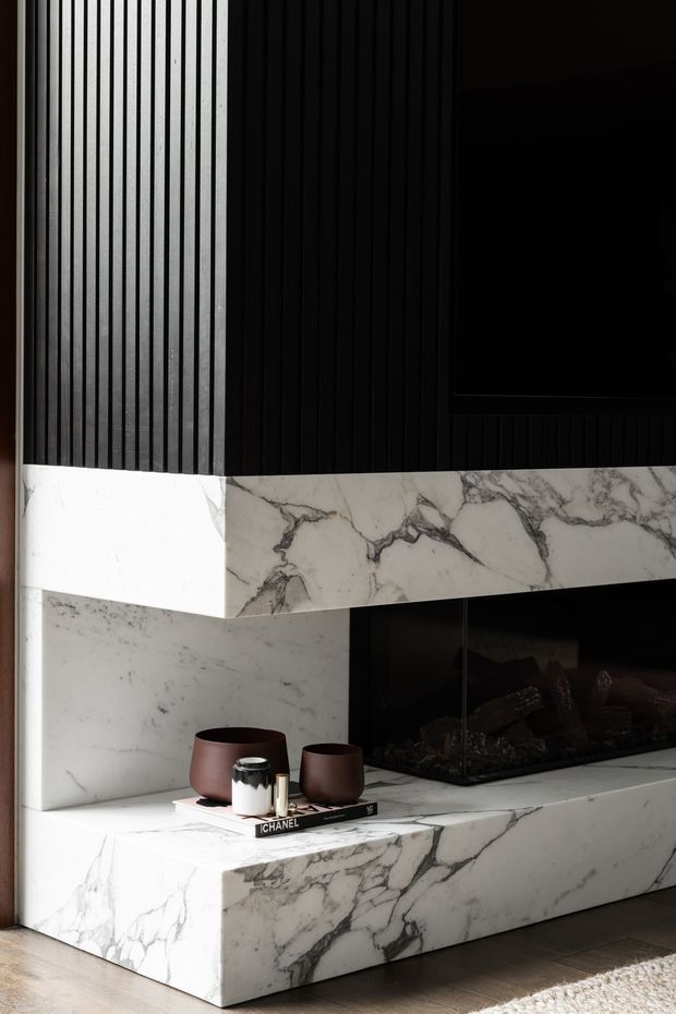 A dramatic contemporary living room fireplace feature clad in richly veined white marble. The composition includes a long marble hearth and mantel, a black inset fireplace, dark vertical ribbed wall panelling, and a black television above. Small decorative vessels and books sit on the hearth, with timber flooring and a rug visible below.