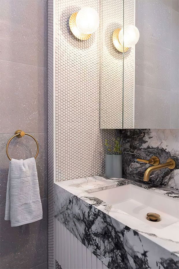A refined contemporary bathroom featuring a wall-mounted basin with a dramatic white-and-black veined stone vanity and splashback. The walls combine large grey tiles with a small round mosaic tile feature, complemented by brushed brass wall-mounted tapware, a circular towel rail, and globe wall lights. A mirrored cabinet and a small planter add functional and decorative detail.