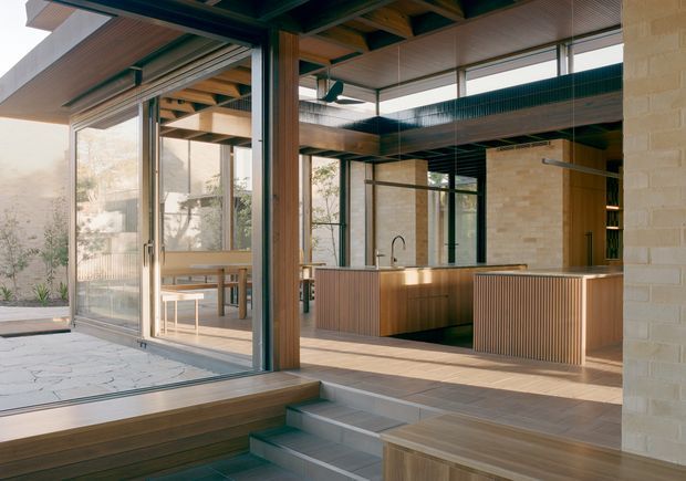 A contemporary open-plan kitchen and dining area connected to a covered outdoor terrace through expansive sliding glass doors. The kitchen features warm vertical timber cabinetry, a large island with an integrated sink and dark benchtop, pale brick walls, exposed timber ceiling structure, dark metal-framed glazing, and stepped levels leading toward a lower seating or outdoor area. The space is flooded with natural light and has a warm, indoor-outdoor character.