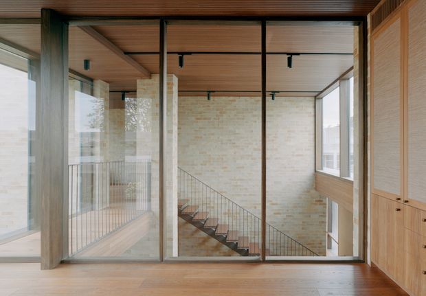 Interior view of a contemporary stairwell and landing, framed by large black metal and glass partitions. A timber-lined ceiling and warm timber flooring complement pale exposed brick walls, while a slim metal stair balustrade descends through the central double-height space. Built-in timber cabinetry occupies the right side, and extensive glazing brings in natural light.