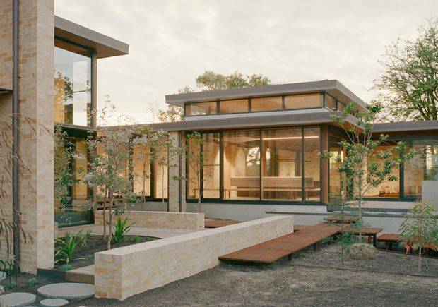 Exterior view of a low-rise contemporary residence arranged around a landscaped courtyard. The building features extensive floor-to-ceiling glazing, pale masonry walls, dark slim window frames, broad low-pitched roof overhangs, and warm timber-lined interiors visible through the glass. A raised timber walkway crosses the courtyard, with built-in pale masonry planter and retaining walls and sparse architectural planting.