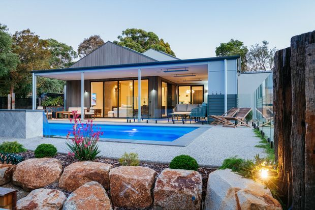 Contemporary single-storey residence viewed from a landscaped garden, with a broad flat-roofed entertaining pavilion, floor-to-ceiling glazed living spaces, an elevated swimming pool, paved terrace, and a rugged stone retaining wall in the foreground. The home combines dark vertical cladding, pale rendered walls, blue-grey metal detailing, and warm interior lighting.