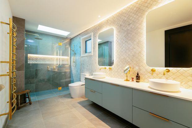 A spacious contemporary bathroom with a double vanity, two vessel basins, gold-toned tapware and hardware, rounded-rectangle mirrors, and a large wall-mounted television. The room includes a glass-enclosed shower with blue-grey tiled walls, a freestanding toilet, a skylight, and a distinctive cream patterned wall tile with arched openings. The palette combines pale grey cabinetry, cream and grey tiles, and warm metallic accents.