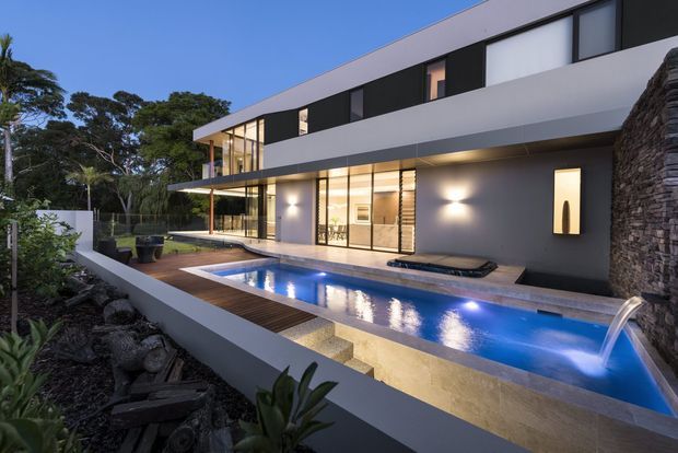 Exterior dusk view of a large two-storey contemporary residence with strong horizontal lines, extensive floor-to-ceiling glazing, projecting upper-level volumes and a cantilevered roof or awning. A long illuminated swimming pool with a water feature occupies the foreground, bordered by timber decking and pale paving. Dark stacked-stone walling and landscaped garden beds frame the property.