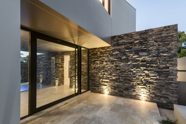 Contemporary exterior courtyard or terrace beside a modern home, featuring large black-framed glazed doors, a cantilevered upper volume, pale stone-look paving, and a dramatic layered dark stone feature wall illuminated from below.
