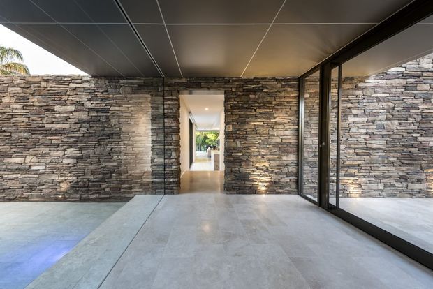 A contemporary covered entry or transitional space with extensive stacked natural-stone walls, large-format pale stone flooring, a dark flush-panel ceiling, and full-height black-framed glazing. The glazing reflects the stone wall and opens toward a bright interior corridor and landscaped exterior beyond.