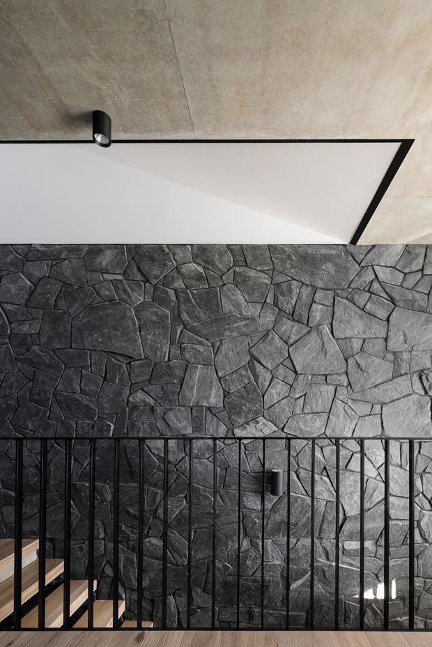 Interior architectural detail showing a black metal stair balustrade in front of a dramatic irregular dark stone feature wall. A timber stair is partially visible at lower left, while the ceiling combines pale textured concrete or plaster with a sharply angled white soffit edged in black. The composition is monochromatic, tactile, and contemporary, with a single black cylindrical spotlight.