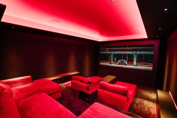 A dark, high-end home cinema with tiered red upholstered lounge seating arranged around a central coffee table. The room has dark wall and floor finishes, a large projection screen or glazed display at the front, and dramatic red cove lighting across the ceiling and along the walls, creating an immersive theatrical atmosphere.
