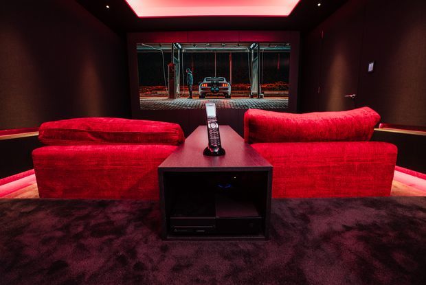 A dark, luxurious home cinema viewed from behind two vivid red sofas facing a large projection screen. The room features black walls, a deep plush carpet, a central media console with remote control, concealed low-level lighting, and red illuminated ceiling detailing. The screen displays a garage-like scene.