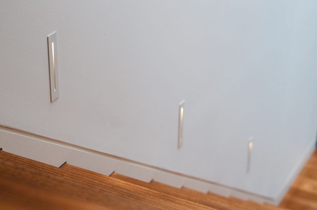 Close-up view looking down a narrow staircase with warm timber treads, white painted walls, white skirting boards, and slim vertical wall-mounted light fixtures.