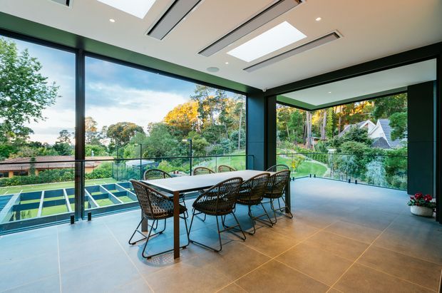 A spacious, contemporary enclosed outdoor entertaining area with floor-to-ceiling glazing, black-framed windows, skylights, and a large dining table surrounded by woven chairs. The room opens visually to a landscaped suburban setting and overlooks a sports court or pool-like recreational area. Large-format warm grey floor tiles and dark structural framing create a refined indoor-outdoor atmosphere.