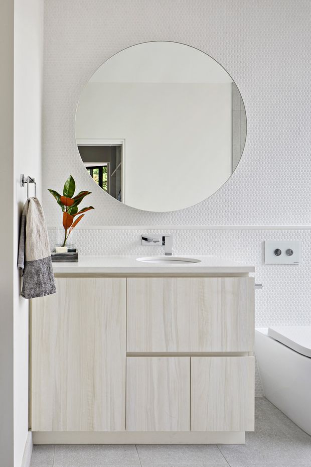 Bright contemporary bathroom with a pale timber floating vanity, integrated white basin and chrome tapware. The walls are clad in small white textured mosaic tiles with a decorative horizontal trim. A large round frameless mirror dominates the wall above the vanity, while a freestanding white bathtub is partially visible at right. Neutral grey flooring, a wall-mounted dual flush plate, and a small plant and hanging towels complete the clean, refined space.