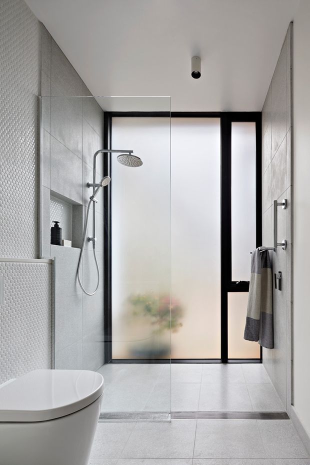 Contemporary bathroom with a walk-in glass shower, wall-mounted rainfall and handheld shower fixtures, pale grey tiled walls and floor, textured feature tiles, a sculptural white toilet, and black-framed frosted glazing. The room has a clean, restrained and softly illuminated aesthetic.