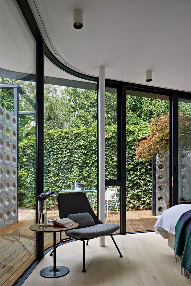 A bright, refined bedroom or reading retreat with floor-to-ceiling curved glazing in black frames, opening onto a timber deck and lush garden. A sculptural grey lounge chair and small round side table sit on a pale timber floor beside a partially visible bed. White cylindrical columns, minimalist ceiling lights, and distinctive perforated concrete block screens frame the space.
