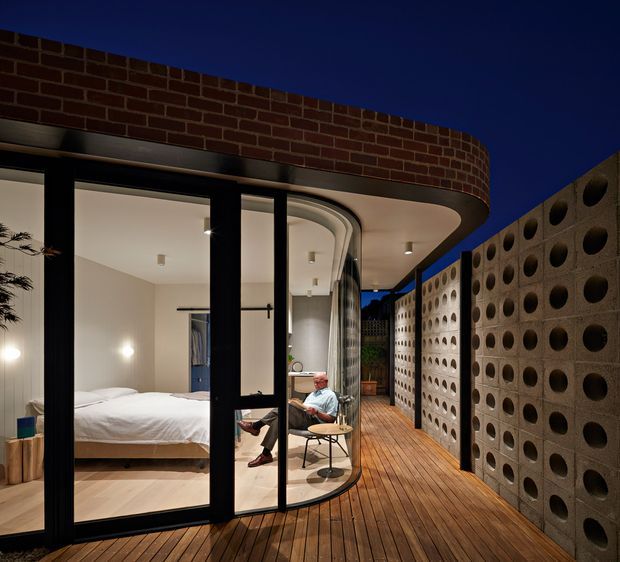 A modern residential courtyard or terrace at dusk, featuring a curved glazed bedroom pavilion opening onto a timber deck. The bedroom has minimal white bedding and pale walls, while a seated person occupies a small chair outside. A curved exposed brick parapet and roof edge frame the upper portion, and a tall perforated concrete breeze-block privacy wall lines the deck, creating a sheltered and textural outdoor space.