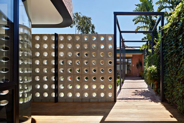 A sunny contemporary courtyard passage beside a curved glazed facade, featuring a perforated concrete breeze-block privacy screen, timber decking, dark steel pergola-like framing, brickwork, and dense green planting along the walkway.