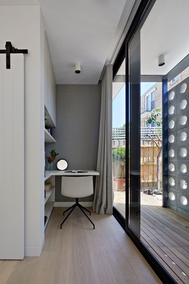 A compact, refined home office nook positioned beside full-height black-framed glazing and a glazed sliding door opening to a timber deck and small courtyard. The room features pale timber flooring, white built-in shelving and a floating desk, a simple white chair, grey textured feature wall, full-length grey curtains, and minimalist ceiling lights. A perforated concrete-block privacy screen is visible outside.