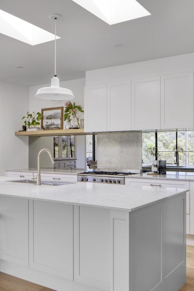 Bright, spacious white kitchen with a large island, shaker-style cabinetry, pale stone benchtops, integrated sink and a professional freestanding range. A textured grey splashback, timber open shelf, pendant light, skylights and windows overlooking greenery add visual interest.