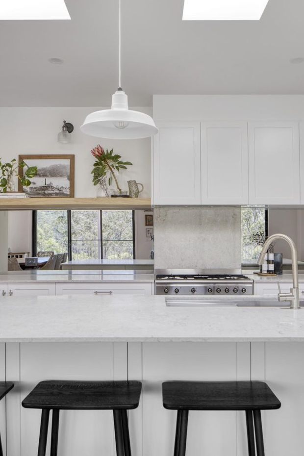 Bright contemporary kitchen with a large pale stone island, white paneled cabinetry, a textured light backsplash, integrated cooktop and sink, black bar stools, black-framed windows, and a white pendant light. A timber shelf displays framed artwork and plants above the rear work area.