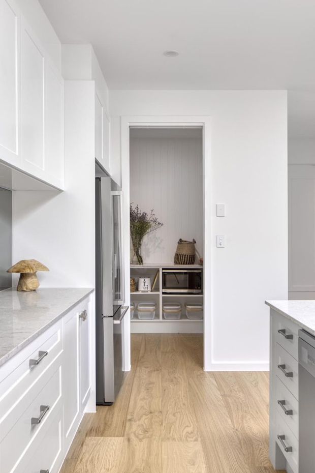Bright white kitchen with shaker-style cabinetry, pale stone benchtops, stainless-steel appliances, and a light timber floor. The view looks toward a compact walk-in pantry with open shelving, drawers, a microwave, and vertical wall panelling. The space has a clean, refined, understated appearance with abundant white surfaces and natural wood tones.