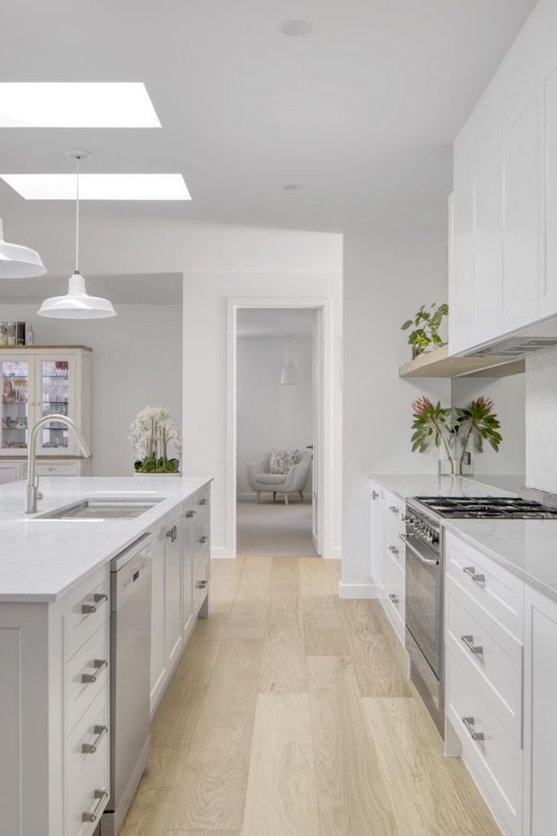 Bright, narrow galley-style kitchen with white Shaker cabinetry, pale engineered timber flooring, light stone benchtops, stainless-steel appliances, and a central island with sink. Skylights and pendant lights provide abundant natural light. A doorway at the rear leads to a softly furnished living area, while plants and glass-fronted storage add warmth.