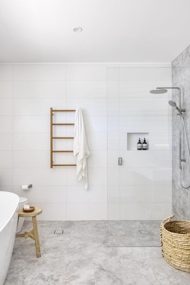 Bright contemporary bathroom featuring large white wall tiles, a grey stone-look tiled floor, a walk-in shower with dual showerheads and recessed niche, a freestanding bathtub edge, and a warm timber towel ladder. A woven basket and small timber stool add natural texture.