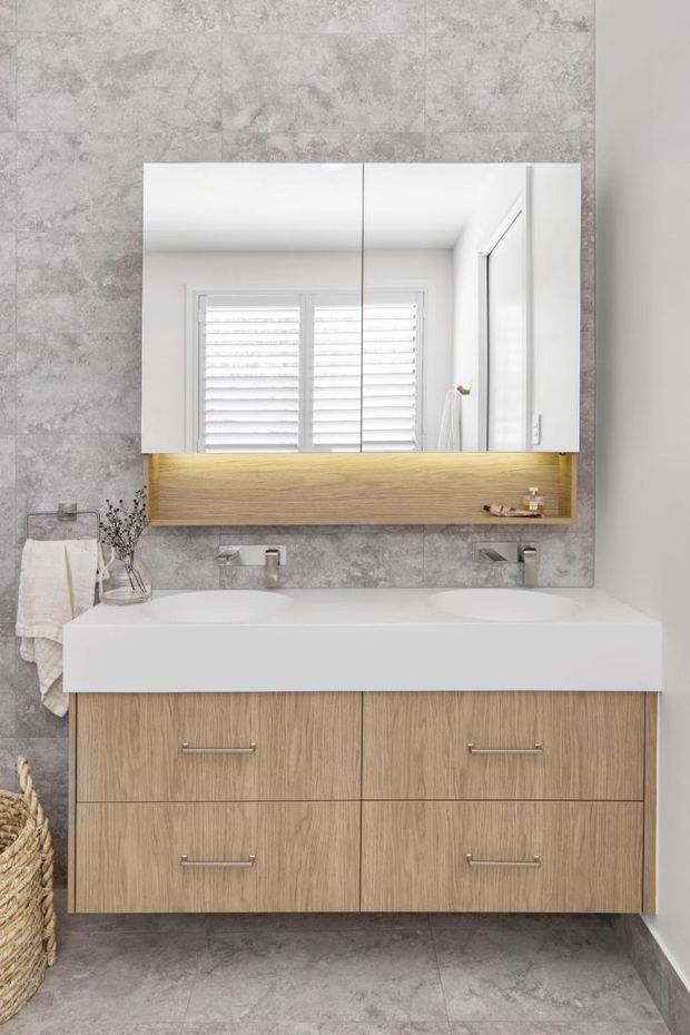Bright contemporary bathroom featuring a wall-mounted double vanity with a white integrated countertop and twin basins, oak-toned drawer cabinetry, a large mirrored cabinet with warm underlighting, grey stone-look wall and floor tiles, and white plantation shutters reflected in the mirror.