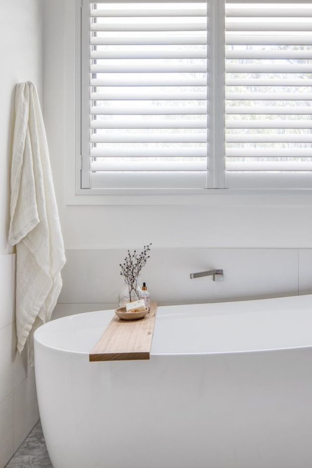A bright, minimalist bathroom featuring a freestanding white bathtub beneath a large white-framed shutter window. A timber bath tray spans the tub and holds a small bowl and decorative items. White wall panelling, a wall-mounted brushed metal tap, a hanging white towel, and grey patterned floor tiles complete the calm, monochromatic space.