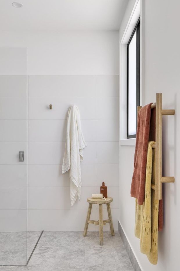 Bright minimalist bathroom with a frameless glass shower screen, large white wall tiles, pale grey stone-look floor tiles, and a black-framed window. A timber ladder towel rack holds terracotta and mustard towels, with a small wooden stool and toiletries adding warm accents.