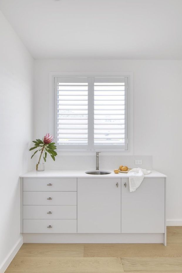 Bright, minimalist white kitchen nook with a compact built-in cabinet run, integrated drawers and cupboards, a white benchtop, and a small undermount sink beneath a white plantation-shutter window. Pale timber flooring adds warmth, while a simple vase with greenery and fruit provide restrained accents.