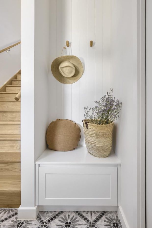 A bright, narrow entryway features a built-in white paneled bench and recessed display niche with hooks, a straw hat, woven basket, and neutral cushion. Decorative patterned floor tiles add contrast, while a timber staircase with a simple handrail is visible alongside the alcove.