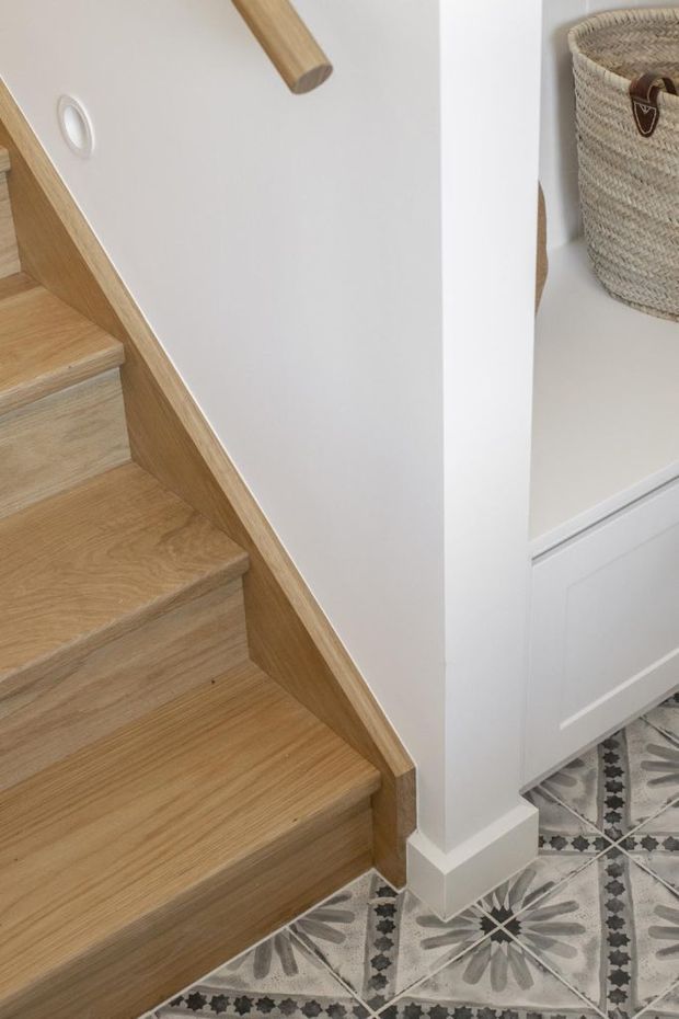 Close-up interior view of a compact stairway with light timber steps, matching timber handrail and side trim, white walls, and integrated white cabinetry beneath the stairs. A woven basket sits in an open shelf, while patterned decorative floor tiles create a contrasting base.